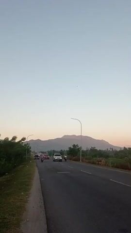The video shows a two-lane road with cars and motorcycles driving on it. There is a grassy shoulder on the left side of the road, with trees and bushes lining the edge. In the distance, there is a large mountain. The sky is a clear light blue, suggesting it is daytime. The overall impression is a peaceful rural scene.
