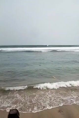 The video shows a choppy ocean with waves breaking near a sandy beach. The sky is overcast and grey. There are several dark rocks in the water, some of which are partially submerged by the waves. The water is a deep blue-grey color and the waves are white-capped. The beach is made of light brown sand. The video is shot from a slightly elevated position, looking out over the water.