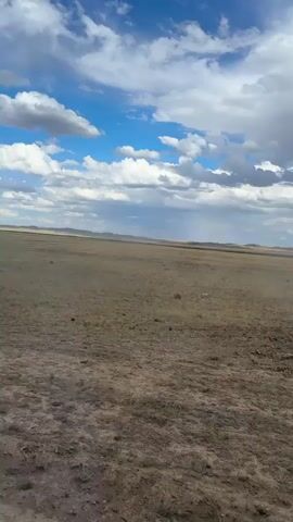 This media file captures a wide-angle, horizontal view of a vast, flat, arid landscape under a dynamic sky. The foreground is a dry, brown, somewhat barren terrain with patches of sparse, dry grass and darker soil. The middle ground extends to a seemingly endless horizon, dominated by a flat, muted brown plain. In the far distance, low, rolling hills or mountains are faintly visible, their outlines softened by atmospheric haze.

The sky is a prominent feature, filled with a dramatic mix of bright blue patches and large, fluffy white and gray clouds. These clouds suggest a day with changing weather, possibly with a storm brewing or passing in the distance, as indicated by faint vertical streaks of rain or mist in the far background. The lighting suggests it is daytime, likely mid-afternoon, given the brightness of the blue sky and the shadows cast by the clouds.

There are no visible people or man-made objects in the frame, emphasizing the natural and remote quality of the scene. The overall impression is one of openness, solitude, and the raw beauty of a dry, expansive environment. The location cues, combined with the description of a vast, arid plain and the specific context of Bakty, Kazakhstan, point to a rural or wilderness setting in Central Asia. The mood is serene yet somewhat somber due to the vastness and the overcast sky. There is no visible text in the media file.