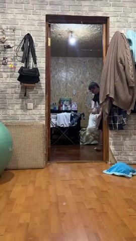 A young man picks up a baby and carries them into a room with a brick-patterned wall, wooden floor, and doorway. The man is wearing a black shirt, black shorts, white socks, and glasses. The baby is wearing a light-colored outfit. The doorway leads into a smaller room with plywood walls, a bed, and various objects on a table. There is also a green exercise ball, a woven mat, a black purse, and some clothing hanging on the wall. The man sets the baby down and appears to be helping them to walk. The baby makes sounds, possibly babbling or attempting to communicate.