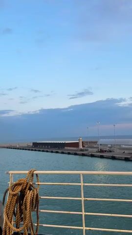 The video shows a view from a ferry or ship, looking towards a pier with a lighthouse and a long building. The sky is partly cloudy with a blue gradient, suggesting either sunrise or sunset. A thick, coiled rope hangs from the railing of the ferry. The water is relatively calm. There is no visible interaction between people or any other significant action occurring in the scene. The pier has street lamps and some vehicles are parked on it.