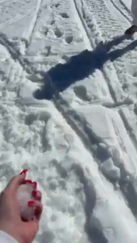 Two people in white clothing are in a snowy area. One person holds a snowball in their hand, and the other person is standing in the distance. The person with the snowball throws it. The camera follows the snowball as it flies through the air and lands on the ground.
