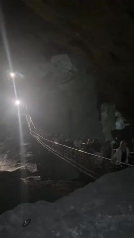 A group of people are walking up a metal ramp in a cave. The ramp is lit by several spotlights, which cast long shadows. The cave walls are rough and rocky. The people are dressed in casual clothing. Some are holding their phones up, possibly to take pictures. The overall atmosphere is dark and mysterious.