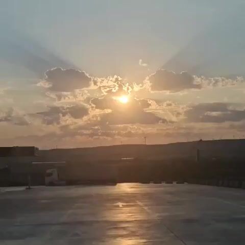 A video displays a wide shot of a cloudy sunset with the sun peeking through the clouds. The sun's rays are visible, fanning out from behind the clouds. In the foreground, there is a flat, paved surface, possibly a road or parking lot, reflecting the light of the sunset. To the left, a white semi-truck is parked. In the background, there are hills or a slightly elevated landscape with some structures and what appear to be wind turbines on the horizon. The overall scene is one of a natural landscape transitioning into evening.