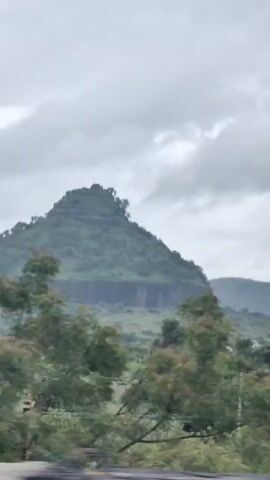 This is a vertical, shaky video clip featuring a large, green, conical hill with a flat, stepped plateau at its summit. The hill has a sheer, dark, rocky base. The surrounding landscape consists of rolling green hills, scattered trees, and what appears to be agricultural fields. A white building and some power lines are visible in the mid-ground. The sky is overcast with dramatic, grey clouds. The camera is in motion, suggesting it's mounted on a vehicle moving past the scene.