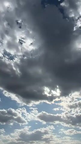The video shows a time lapse of clouds moving across a blue sky. The clouds range in color from white to dark gray, indicating varying thickness and density. Sunlight filters through some of the clouds, creating bright patches and shafts of light. The camera is pointed upwards, capturing the dynamic movement of the sky. There are no people or objects visible in the frame.