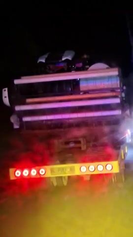 A low-angle rear view of a large truck or trailer moving at night is shown. The vehicle's cargo is illuminated by multiple horizontal tube lights that cycle through red, blue, and purple hues. On top of the cargo, the silhouette of a smaller vehicle, resembling an ATV, is visible. The rear bumper features a horizontal bar with several circular red lights and a yellow center section displaying the license plate "BL 7088 H". A hazy cloud, possibly exhaust or dust, is illuminated by the vehicle's lights near the ground. A siren sound is continuously audible throughout the video.