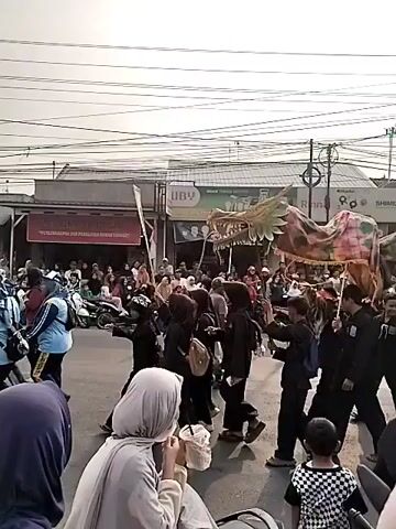 A parade is taking place on a street lined with shops. People are walking along the street, and some are watching the parade from the sidewalks. The parade features a large dragon costume being carried by several people. The dragon is colorful and appears to be made of fabric. Some participants are wearing black uniforms, while others are in casual clothing. A few motorcycles are visible in the background, suggesting this is a public street. The sky is overcast, and there are power lines overhead. The overall atmosphere seems festive.