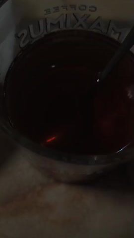 The video shows a close-up of food and drink. In the first frame, a glass of dark liquid, possibly tea or coffee, is visible with a spoon inside. The glass has writing on it. The subsequent frames focus on a white plate filled with numerous dumplings. The dumplings are light brown and appear to be boiled. A fork is partially visible in the background, resting on the plate. The lighting is dim, suggesting it might be evening or indoors. The overall scene depicts a simple meal.
