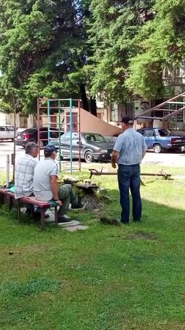 The video shows a group of three men outdoors in a grassy area with trees and buildings in the background. Two men are sitting on a bench, facing away from the camera, while the third man stands facing them, holding a phone. There is a chess set on the ground between the bench and the standing man, with chess pieces visible. There are cars parked in the background, and playground equipment is visible behind the bench. The scene appears to be a park or recreational area.
