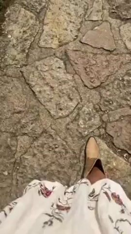 The video starts with a close-up of someone walking on a stone path, wearing cowboy boots and a short white skirt. The camera then pans up to show a group of people walking along the path, with mountains in the background. The camera pans up further to reveal a lake surrounded by mountains and green fields. A winding road follows the shoreline, and the sky is partly cloudy.