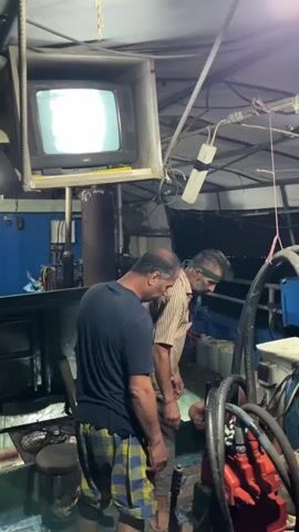 The video shows three men inside what appears to be the engine room of a boat. A CRT television is mounted overhead and displays a bright, washed-out image. One man, wearing a dark blue t-shirt and plaid shorts, stands looking down. Another man, wearing a striped shirt and a green band around his head, also looks down. A third man, shirtless and with dark shorts, is partially visible in the background, appearing to be working on something. The environment is cluttered with mechanical equipment, hoses, and storage containers. The lighting is dim, suggesting it is either nighttime or the interior is poorly lit.