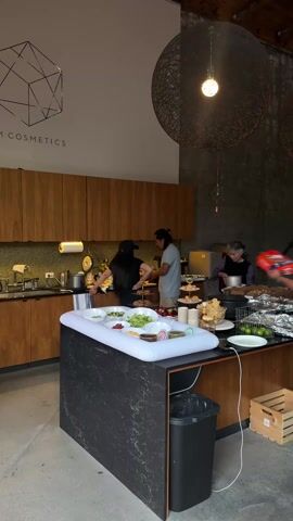 The video shows a bright, open indoor space with several people present. In the foreground, an island countertop is visible, filled with various food items, including salads and snacks. People are gathered around the island, seemingly preparing or serving food. The kitchen area with wooden cabinets is visible in the background. To the right of the island, there is a table with more food items covered in foil. As the camera pans right, more people are seen seated at another table, with a red bag and a box of snacks nearby. The room has a high ceiling with large, decorative light fixtures, and a staircase is visible in the background. The walls are a mix of painted surfaces and exposed brick. The overall atmosphere suggests a casual gathering or event where food is being prepared and shared.