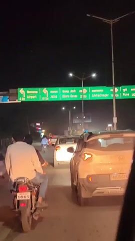 The video captures a nighttime scene on a road, likely in India, with multiple vehicles visible. A green directional sign with white text is prominently displayed overhead, indicating distances and directions to various locations, including "Airport" (6 Km), "Jalna" (61 Km), "Ellora Caves" (33 Km), and "Ahmednagar" (118 Km). Below this, a smaller blue sign reiterates some of these directions.

In the foreground, a person on a motorcycle, wearing a white shirt, is seen from behind. The motorcycle has a license plate starting with "MH 20 GE 9965". Beside it, a white car is stopped in traffic, its rear visible, and its license plate reads "MH 20 GE 4285". Other vehicles are present in the background, suggesting a traffic jam. The environment is dark, illuminated by streetlights and the headlights of the vehicles. The overall impression is of a typical roadside scene at night with traffic moving towards several destinations.