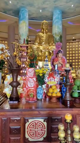 The scene features a richly adorned altar, likely within a Buddhist temple. At the center, a large, gleaming golden Buddha statue sits atop the altar. Flanking the Buddha are two painted pillars, each depicting a mountain landscape with a cloudy sky. In front of the Buddha, a smaller statue of a child-like figure sits cross-legged in a posture of prayer. 

The altar itself is made of dark wood with intricate carvings. It displays an array of offerings including vases of yellow flowers, stacks of red gift boxes, oranges, and green pomelos. A small pagoda-like structure adds to the visual complexity. Candles in ornate holders are placed on either side of the altar. The backdrop is a wall of small, rectangular cells, each potentially holding a votive offering or similar symbolic item. The ceiling above is decorated with a painted sky and indirect lighting that enhances the serene and spiritual atmosphere of the setting.