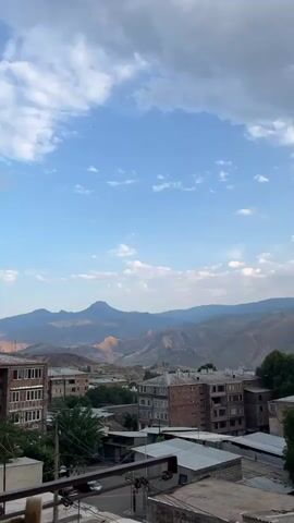 This is a vertical, wide-angle video shot from a high vantage point, looking out over a town nestled against a backdrop of rolling, barren mountains under a cloudy sky. The scene appears to be an outdoor urban setting in the daytime, likely late afternoon given the soft light.

In the foreground, the tops of several buildings are visible. These are mostly low-rise structures with flat, metal roofs, and some appear to be older concrete apartment buildings with visible balconies. Lush green trees are interspersed among the buildings, suggesting a moderate amount of vegetation within the town. A partial view of a balcony railing is present at the bottom left of the frame.

The midground features more buildings of varying architectural styles, including some taller, multi-story apartment blocks constructed from what looks like concrete and brick. The town appears to be built on a slope, leading up towards the mountains.

The background is dominated by a vast expanse of mountains. These mountains are layered and appear arid, with their slopes showing shades of brown, beige, and muted green. A prominent, solitary peak stands out in the far distance, adding a dramatic element to the landscape.

The sky is a mix of blue patches and large, dramatic clouds. Some clouds are white and wispy, while others are darker and more voluminous, suggesting potential for weather changes. Tiny dark specks, possibly birds or insects, are visible scattered across the sky.

The overall mood is serene and somewhat contemplative, with the natural grandeur of the mountains contrasting with the man-made structures of the town. The time of day appears to be afternoon, and the weather is partly cloudy. The location is identified as Gladzor, Armenia, which aligns with the mountainous terrain and architectural style. No text is visible in the video.