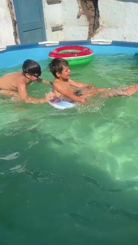 In a bright, outdoor setting, two young boys enjoy a day in a shallow swimming pool. The pool water is a vibrant, clear green, reflecting the sunny day. The larger boy, wearing black goggles, is in the foreground, his arms extended as if supporting or pushing the other boy. He is wearing black swim trunks. The younger boy, on the right, is seated on a translucent inflatable ring, his legs dangling in the water. He is looking to his right. A red and green inflatable watermelon float is visible to the left of the boys, partially submerged. In the background, a yellow inflatable toy, possibly a duck, bobs near the edge of the pool. Behind the pool, a white wall with some texture and a blue door suggest a backyard or patio area. The sunlight is strong, casting dappled shadows on the water and illuminating the scene. The overall impression is one of playful summer fun.