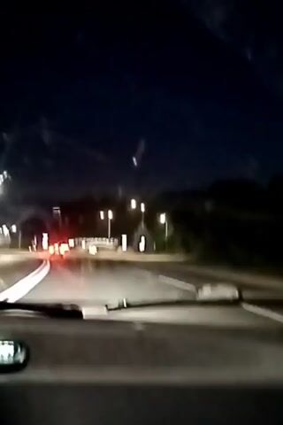 The video shows a nighttime view from inside a car driving on a road. The sky is dark blue. Streetlights are visible along the side of the road, illuminating trees and white signs with dark text. A car with red taillights is ahead on the road, appearing to turn. The road surface is wet, reflecting the lights. The car's windshield wipers are visible in the foreground. The sign in the distance reads "ЗЕЛЕНОдольск" in large, illuminated letters.