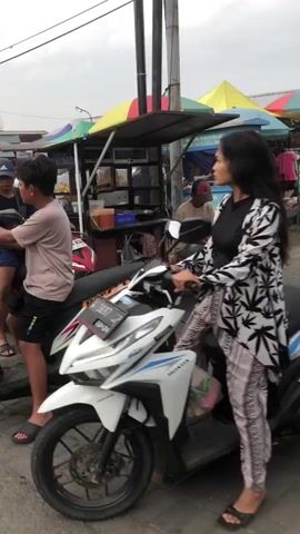 The video shows a street scene, likely in a Southeast Asian country, with several people and a food stall. In the foreground, a white Honda scooter is parked, with a young woman standing next to it, holding the handlebars. She is wearing a black top and a patterned white and black cardigan, with matching patterned pants and black sandals. Her right leg is resting on the footrest of the scooter. To the left of the scooter, a young man is seated on another motorcycle, wearing a light brown t-shirt and black shorts. He is looking towards the right. Behind them, a food stall with a colorful awning displays various food items. A man in a red cap is visible behind the counter of the stall. The ground is paved with interlocking bricks. The overall atmosphere appears to be casual and busy.