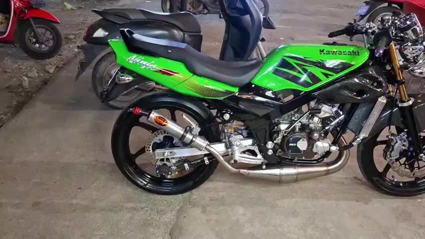 This media file features a vibrant green and black Kawasaki Ninja motorcycle parked on a concrete surface, likely outdoors. The motorcycle is the primary subject, displayed in profile view, showcasing its sporty design and modifications. It has a sleek, aerodynamic fairing, a custom exhaust system with an orange end cap, and black alloy wheels. The "Ninja" logo is visible on the tail section, and "Kawasaki" is emblazoned on the fuel tank.

In the background, other motorcycles are partially visible, suggesting a parking area or a gathering of vehicles. A red scooter is parked to the left, and a black scooter or motorcycle is behind the Ninja. The setting appears to be an outdoor space with a textured concrete floor. The lighting suggests it might be daytime, possibly late afternoon or early evening, as there are no harsh shadows and the light seems diffused. There are hints of red chairs in the far right background, suggesting a nearby seating area or establishment.

No people are clearly visible or actively engaged in any activity directly related to the motorcycle, though their presence might be implied by the surrounding vehicles and potential seating. The overall impression is one of presentation or display of a customized motorcycle. The atmosphere seems calm and observational, focusing solely on the visual appeal of the vehicle. No specific event or action is taking place; the media appears to be a static capture or a slow pan of the motorcycle. The context provided indicates the location is Rhieng Blang, Indonesia, which helps contextualize the type of motorcycles and the surrounding environment.