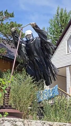 A scarecrow dressed in black plastic trash bags and a white baseball cap is positioned in front of a house. The scarecrow wears a Guy Fawkes mask and holds a broom. The background shows trees, a house with white siding, and a wooden structure. Some plants and a rock wall are visible in the foreground.