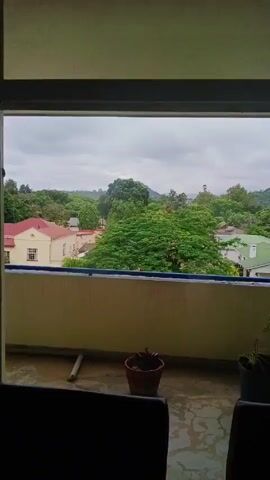 The video shows a view from a balcony overlooking a residential area filled with lush greenery. The sky is overcast. In the foreground, there are two black chairs and a few potted plants on the balcony floor. The balcony has a low wall with a blue trim. Beyond the balcony, there are houses with red and green roofs, interspersed with trees. The scene appears to be calm and quiet.