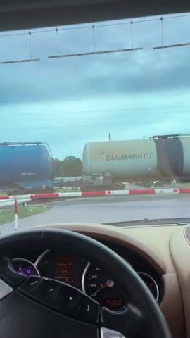 The video shows a driver's point of view from inside a Porsche Cayenne stopped at a railroad crossing. The dashboard is visible, showing a temperature of 15.6°C and the car is stationary (0 km/h). Through the windshield, railroad tanker cars labeled "BULMARKET" are visible on the tracks. The crossing arms are down. As the camera pans to the left, the side mirror captures more of the tanker cars and the railroad crossing signal.