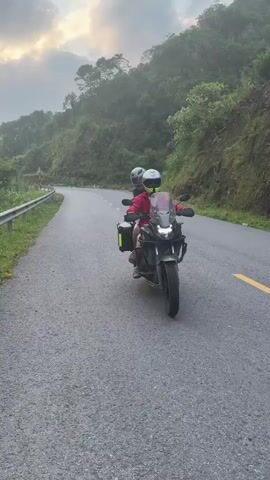 The video shows two people on a black Honda CB500X motorcycle, stopped on a paved, winding road in a mountainous region. The background features lush, green, forested hills, partially obscured by haze or mist under an overcast sky, suggesting early morning, late afternoon, or cloudy weather. The location is Tân Quang Commune, Vietnam.

The driver is wearing a bright red jacket over a white shirt, light-colored shorts, and sandals. They have a white and neon yellow full-face helmet with "YOHE" visible on the side, sunglasses, and a camera mounted on the chin of the helmet. Text on the red jacket partially reads "riders" and "ball." The motorcycle's handguards display "S.WOLF." The passenger, seated behind the driver, wears a dark top, a grey full-face helmet, and dark pants or shorts.

The Honda CB500X motorcycle is equipped with black hard-shell luggage boxes on both sides of the rear, one featuring a neon yellow stripe. "HONDA" is visible on the engine casing. The road surface is asphalt with clear yellow painted lines. The riders appear to be taking a break, looking around the scenic surroundings and conversing, indicating a relaxed pause during a journey through the rugged landscape.