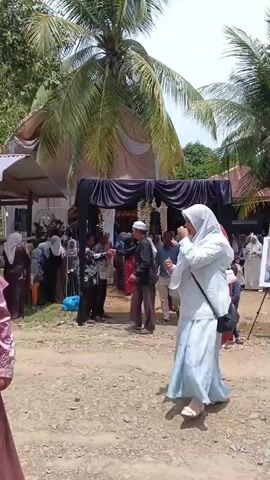 The video shows an outdoor gathering, likely a ceremony or event, with several people present. In the foreground, a person wearing a light blue hijab and a white top walks away from the camera. Behind them, a group of people are gathered under a tent. To the right, a framed picture of two individuals is displayed on a stand. Coconut trees are visible in the background, suggesting a tropical or subtropical location. The ground is covered in dirt and gravel. Some individuals are seated, while others are standing and conversing. The overall atmosphere appears somber, with black drapery visible near the entrance of the tent.