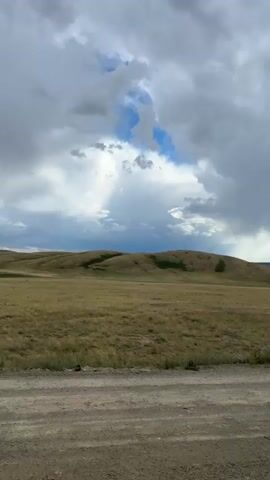 This media file captures a scenic view of a rural landscape in Bakty, Kazakhstan. The scene is dominated by rolling hills covered in dry, golden grass, suggesting a late summer or early autumn setting. In the foreground, a dusty, gravel road stretches across the frame, indicating a remote or undeveloped area.

The sky above is dramatic, filled with a mix of heavy, dark storm clouds and patches of bright blue sky where sunlight breaks through. These sunbeams create a sense of depth and highlight the vastness of the open terrain. The overall weather appears to be transitional, with the possibility of rain or a recent shower in the distance.

There are no people or man-made structures immediately visible, emphasizing the natural and expansive character of the location. The mood is one of quiet solitude and the raw beauty of nature. The time of day is difficult to pinpoint precisely due to the cloud cover, but the quality of light suggests it could be late afternoon or early evening. The rolling hills and the open, grassy plains are characteristic of steppe or prairie environments.

No visible text is present in the media file. The focus is entirely on the natural landscape and the atmospheric conditions.