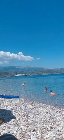 The video shows a sunny day at a beach. The water is clear and blue, and the seabed is covered with pebbles. Several people are in the water, swimming and enjoying themselves. In the background, there are hills and mountains. A large ship is visible in the distance. The sky is a bright blue with a few white clouds.