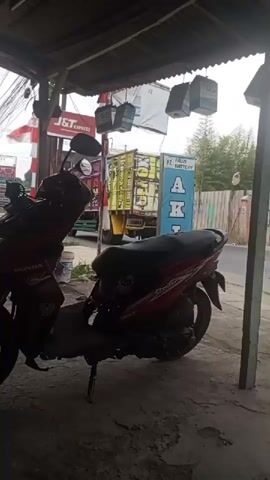 The video pans across a street scene, likely in an urban or semi-urban environment. In the foreground, a red Honda scooter is parked under a makeshift awning, possibly a shop or stall. The scooter is the primary focus of the initial part of the video. Behind the scooter, several items hang from the awning, including what appear to be birdcages and advertisements for J&T Express and GS batteries. A tall, metal-sided truck is seen driving down the street. Further in the background, a motorcycle with a rider is also visible on the road. The overall environment appears somewhat cluttered, with visible wires and graffiti on a fence in the background. The lighting suggests it is daytime, possibly overcast.