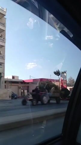 A vehicle drives down a city street on a sunny day. From the passenger seat, the video captures a gas station, other vehicles, buildings, and pedestrians moving past. Solar panels are visible on a building’s roof. The view is partially obscured by reflections on the car window, which is also damaged.