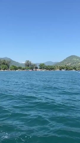 The video shows a view of a beach from the water on a sunny day. The water is blue and has small waves. The beach is sandy and has trees growing along the edge. There are also several boats docked on the beach. In the background, there are green mountains under a clear blue sky.