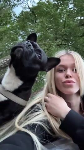 A young woman with long blonde hair and a nose ring smiles at the camera, resting her chin on her hand. Next to her, a black and white French bulldog looks up with its head tilted. They are outdoors, with trees and foliage visible in the background. The woman is wearing a dark hoodie. The dog has a harness on.