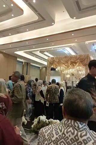 The video shows a wedding reception. There are many people in attendance, dressed in formal attire. The venue appears to be a large ballroom, with high ceilings and ornate decorations. A wedding altar is set up at the far end of the room, adorned with flowers and drapery. Guests are mingling and taking photos. The atmosphere seems festive and celebratory.