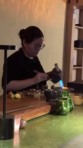 A woman with dark hair in a bun and glasses, wearing a black shirt and apron, stands behind a green countertop preparing food. She uses a small handheld torch with a blue flame to heat a bowl or mortar. Various ingredients and utensils are arranged on the counter, including cut fruits, a wooden cutting board, small jars, and green mugs. Behind her is another person, partially visible, and a shelf holding dishes. The room is illuminated with warm, ambient lighting. The woman is focused on her task, carefully manipulating the torch and ingredients with chopsticks.