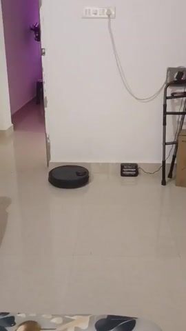 A black robot vacuum cleaner is moving across a tiled floor near a doorway. To the right of the robot, a charging dock for the vacuum is visible, along with a walker. A dark chair is to the left of the robot, and the background shows a hallway with purple lighting.