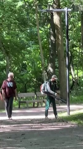 A person in a maroon shirt and black pants walks away from the camera down a dirt path in a park. Behind them, another person wearing green overalls and a baseball cap operates a leaf blower, facing the camera. The leaf blower is a backpack model with an orange gas tank. The person is standing next to a dark gray pole. Further back, a bench and trash can are visible. The area is wooded with green trees and dappled sunlight.