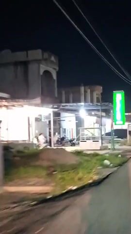 The video shows a nighttime scene outside a building that appears to be a shop or restaurant. A bright green illuminated sign is visible on the right side of the frame. In front of the building, there are several food stalls or carts, some with glass displays. A few people are present, including one person standing near a motorcycle and others sitting or standing under a covered area. A car is parked to the right of the building. The area is dimly lit by overhead lights and the illuminated sign. The camera is moving, suggesting it is being filmed from a vehicle.