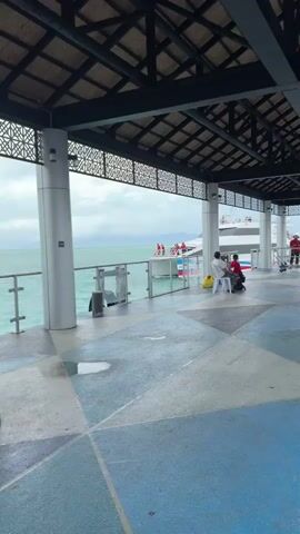 The video presents a view from under the covered area of a pier looking out at a ferry or large boat docked at the pier. The pier has a patterned blue, gray, and white tiled floor and is supported by white pillars. The roof structure above is constructed of dark-colored wood in a geometric design. Outside, the water is a pale green-blue color under an overcast sky. A white and blue boat is docked at the pier, with several crew members wearing red visible on the deck. A banner with the words "HAAD RIN PHANGAN" is displayed on the side of the boat. Two individuals are seated on white plastic chairs near one of the pier's pillars. The person closer to the camera is looking at their phone, while a child is sitting next to them, wearing a red Spiderman hoodie.