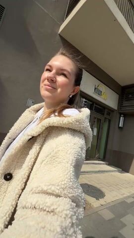 A woman stands outside a modern building and holds the camera up to record herself in selfie mode. The woman has fair skin and brown hair pulled back in a ponytail. She is wearing a white, hooded, fuzzy jacket and a grey zippered sweatshirt underneath. She has small stud earrings. She smiles and looks up at the sun. The building behind her has large glass windows and a stone facade. There are small bushes in front of the building. Traffic sounds are audible.