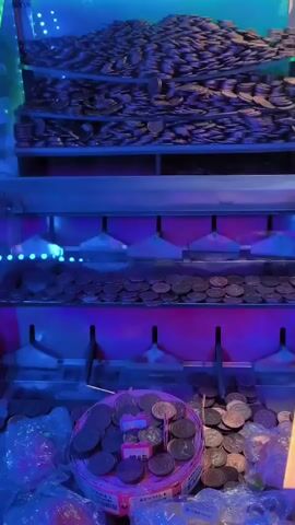 The video shows a close-up of a coin pusher arcade game. The game is illuminated with blue and purple lights. Piles of coins are visible on shelves above and around the main play area. In the center of the play area is a rotating circular dispenser filled with coins. There are also several small plastic bags filled with coins scattered around the bottom of the machine. No people are visible.