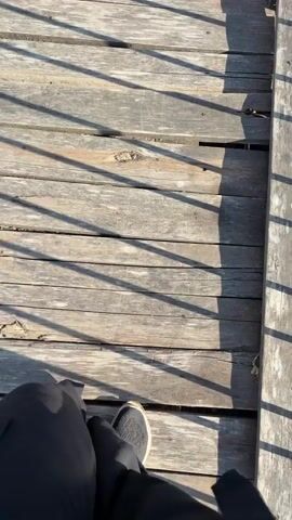 The video shows a first-person perspective of someone walking on a wooden boardwalk. The camera is pointed downwards, focusing on the person's legs and feet, clad in black trousers and a dark athletic shoe. The boardwalk planks are weathered and show visible gaps and wear. Sunlight casts long, distinct shadows from the railing, which runs along the right side of the boardwalk. The railing appears to be made of metal, with some signs of rust and peeling paint. The environment is outdoors, indicated by the natural light and shadows. The person is moving forward, taking steps along the boardwalk.