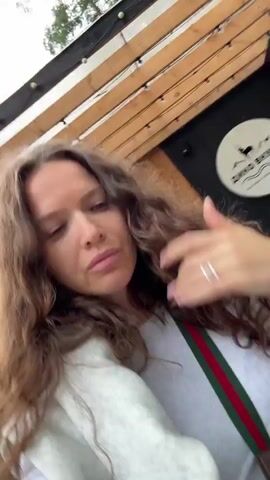 The video shows a woman with long, curly brown hair. She is wearing a white top and a light-colored sweater draped over her shoulders. She is also wearing a necklace with green and red stripes, and a patterned crossbody bag. She has rings on her fingers. She is outdoors in a natural setting with trees and a building visible in the background. The camera appears to be held by the woman, as it is a selfie-style video. The lighting suggests it is daytime.