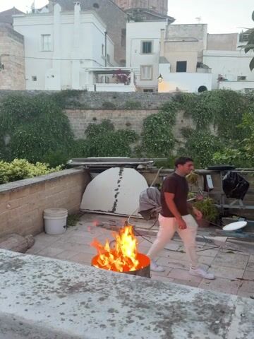 A man in a brown t-shirt and white pants stands to the right of a fire burning in a metal drum. Behind him, a large, white, dome-shaped object is covered with a grey tarp. Further back, a stone wall is covered in green ivy, and white buildings with a tall bell tower rise in the background. The sky is a pale blue.