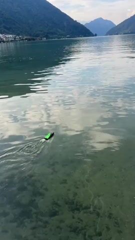 A remote-controlled green boat moves across the surface of a lake, leaving a wake behind it. The lake is surrounded by lush green mountains under a cloudy sky. The water is clear enough to see the sandy bottom.