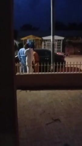 The video shows a dark outdoor scene at dusk or night. In the foreground, there is a wooden structure, possibly a fence or railing. Beyond this, a group of people are visible, their faces indistinct due to the low light and motion blur. They appear to be standing near vehicles and some buildings in the background. A tall pole, likely a utility pole, stands prominently in the middle ground. The sky is a deep blue, indicating it is either early evening or late night. The overall impression is a street or public area, possibly in a town or village.