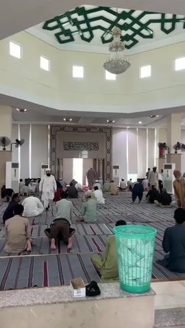 A group of men are gathered inside a mosque, participating in prayer. They are dressed in traditional clothing, with many wearing white or light-colored garments. The men are arranged in rows on prayer mats spread across the floor, facing a decorative wall with Arabic calligraphy. Some men are in a kneeling position, while others are standing or walking. The mosque interior is spacious with high ceilings, decorated with geometric patterns and a large chandelier. Several ceiling fans are visible, and the room is lit by natural light filtering through square windows and artificial lighting. There are air conditioning units and fans mounted on the walls. The overall atmosphere appears to be one of solemnity and community.