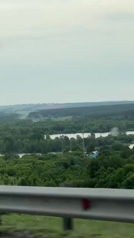 The video shows a wide, flat landscape taken from what appears to be a moving vehicle, with a metal guardrail visible at the bottom of the frame. The scenery is dominated by green trees and a river winding through them, under a cloudy, grey sky. In the distance, cultivated fields can be seen, as well as infrastructure, including a communications tower and some buildings. The focus is on the expansive view, with the landscape stretching towards the horizon.